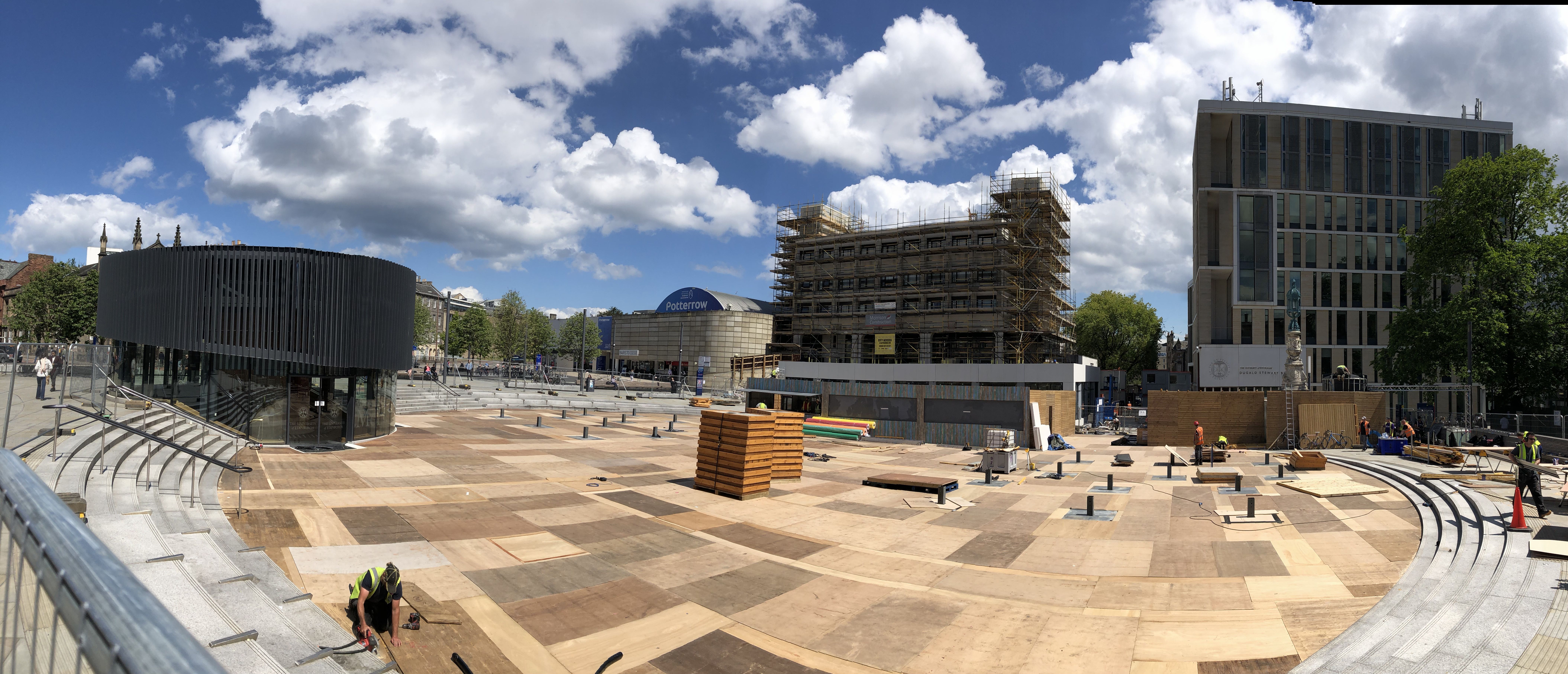 Bristo Square panorama on a sunny day, with wooden boarding being installed across the entire central area, along with small wooden buildings 