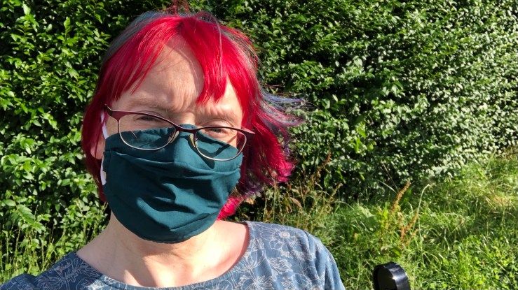 Anna sitting on a park bench during the summer. She is wearing a green face mask, and her hair is vibrant red and blue. There is a green hedge behind her.