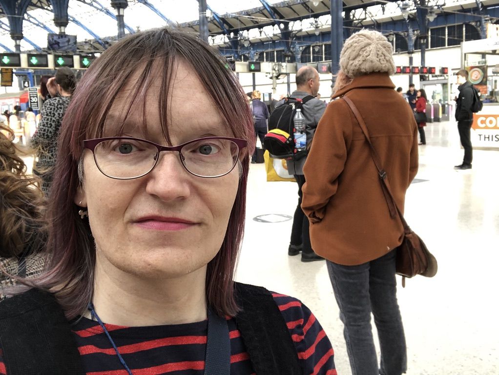 Photo of Anna at Brighton station, wearing a dark pinafore over a red and navy striped top. Her hair is shoulder length with magenta streaks. She is wearing lipstick and has little silver cat earrings. The station is clean and bright and quite busy.