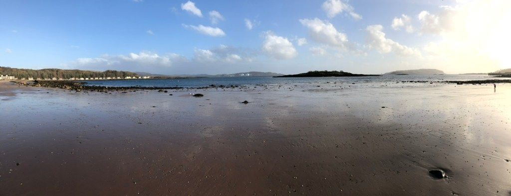 Panoramic photograph of beach on Cumbrae, with the sun low in the sky to the right. The firm wet sand reflects the lightly-clouded blue sky. There is a prominent pebble in the foreground, and a row of houses on the other side of the bay.Small islands are on the horizon.
