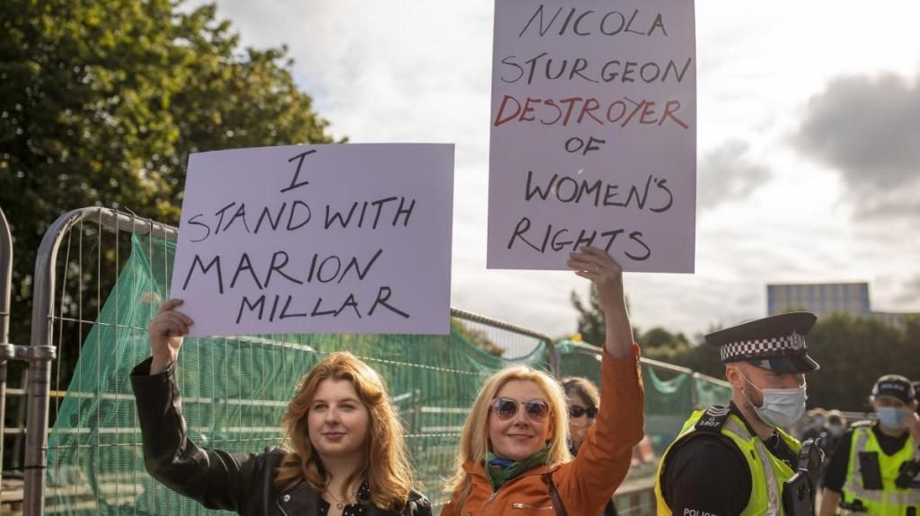 Photo of two unmasked young white women holding hand-written placards aloft, next to a tall metal barrier, with two surgical-face-masked police officers visible nearby. The placards read, ‘I stand with Marion Millar’, and, ‘Nicola Sturgeon, destroyer of women’s rights’.