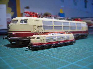 A large electric locomotive in a cream and burgundy livery, modelled in two dramatically different scales. The model in the foreground is less than half height/length of the one behind it..